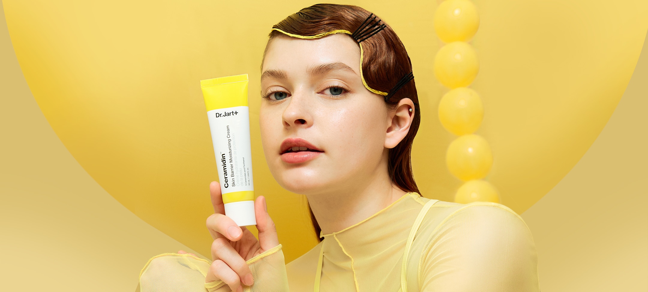 Woman holds tube of Ceramidin Moisturizer Cream in front of yellow abstract background