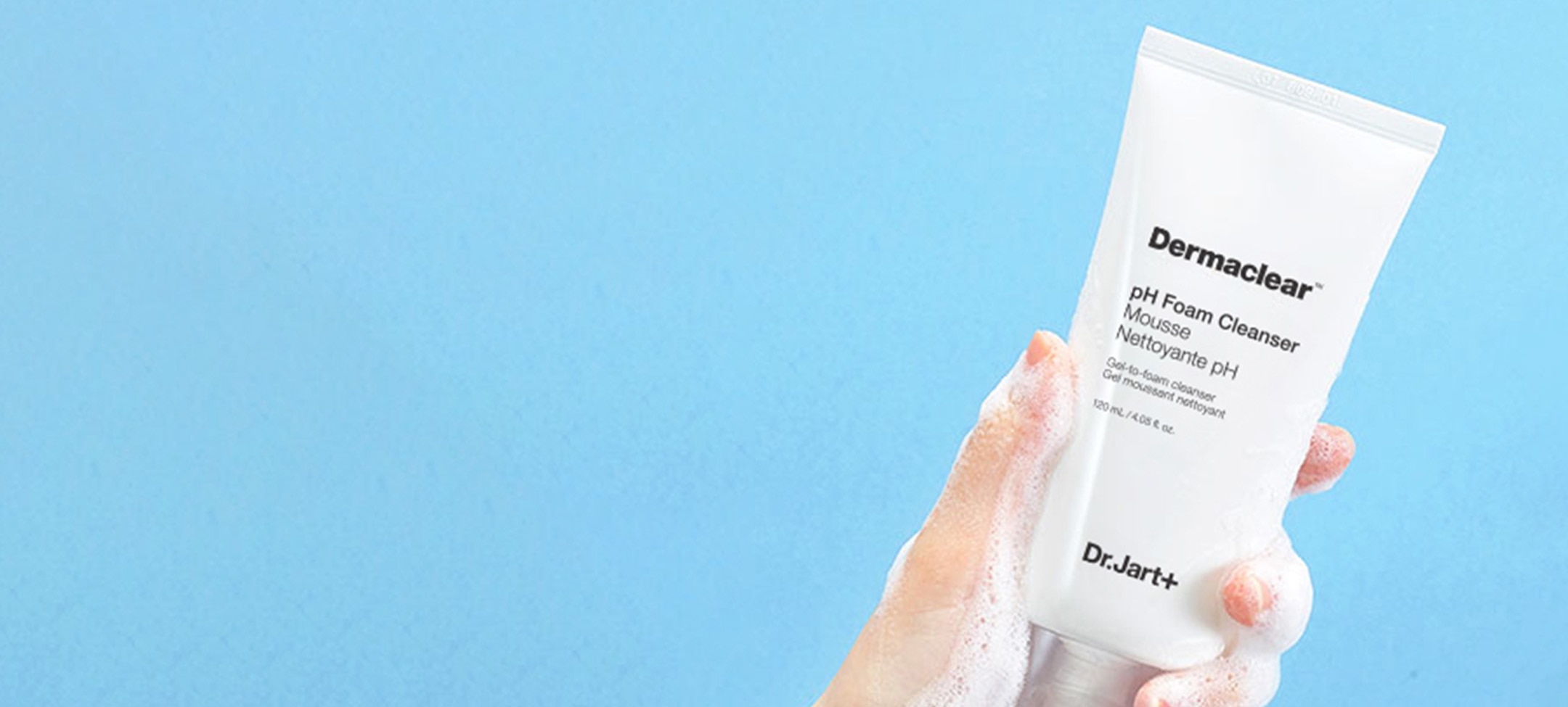 A hand holds up a bottle of Dermaclear Face Wash Cleanser as a frothy, cleanser, face wash flows over the tube and hand. 