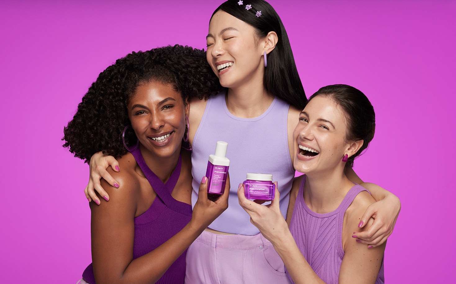 Three young women happily smile & pose holding Prejuvenation Serum & Prejuvenation Cream products in front of a purple backdrop